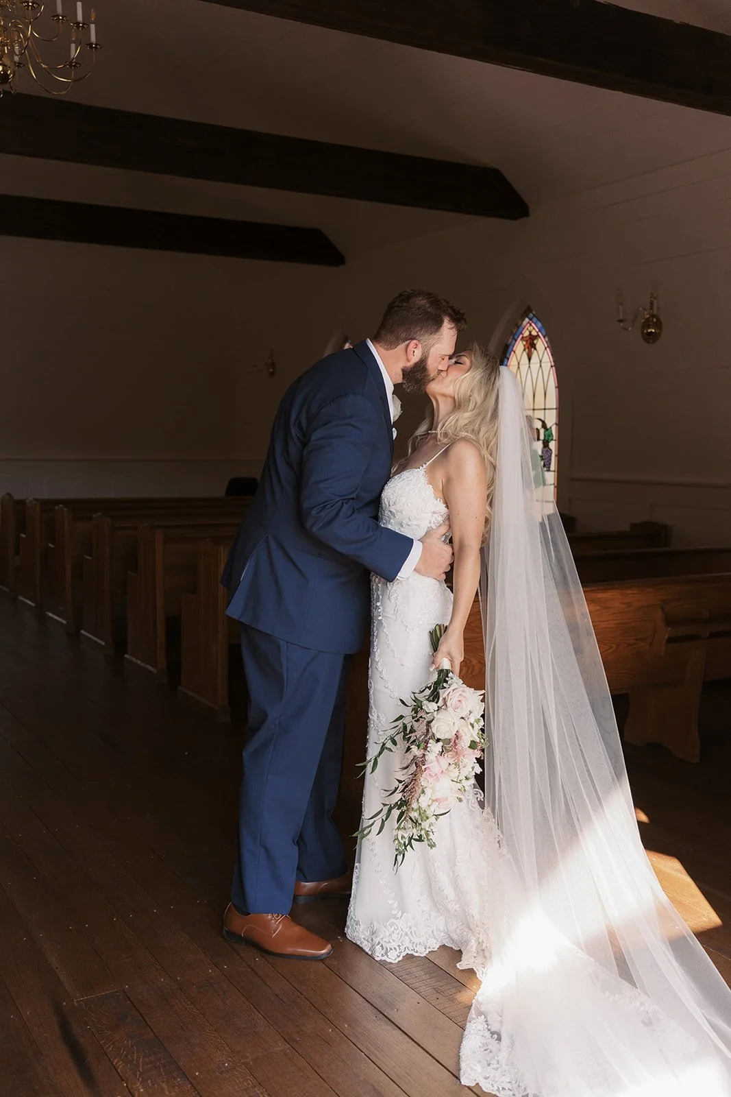 Bride and groom sharing a kiss inside a church, bride holding a bouquet of flowers, stained glass window in the background.