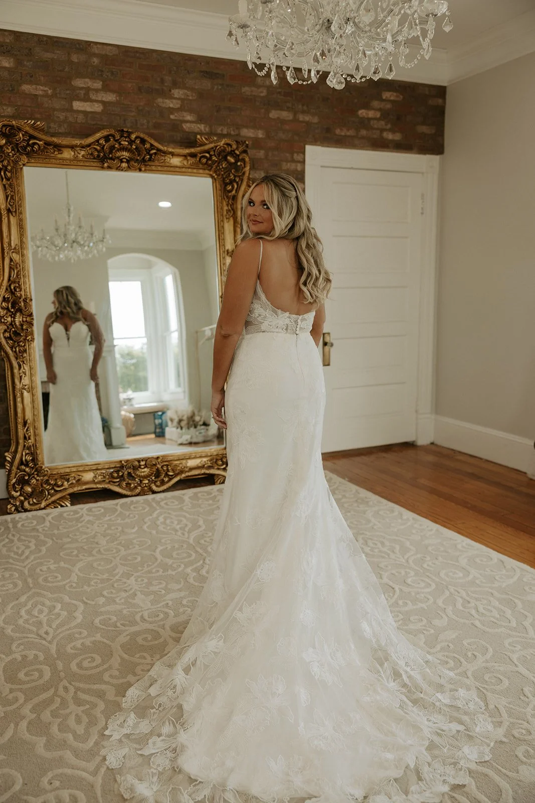 A woman in a wedding dress looking at herself in an ornate gold-framed mirror in a room with a brick wall and a chandelier.