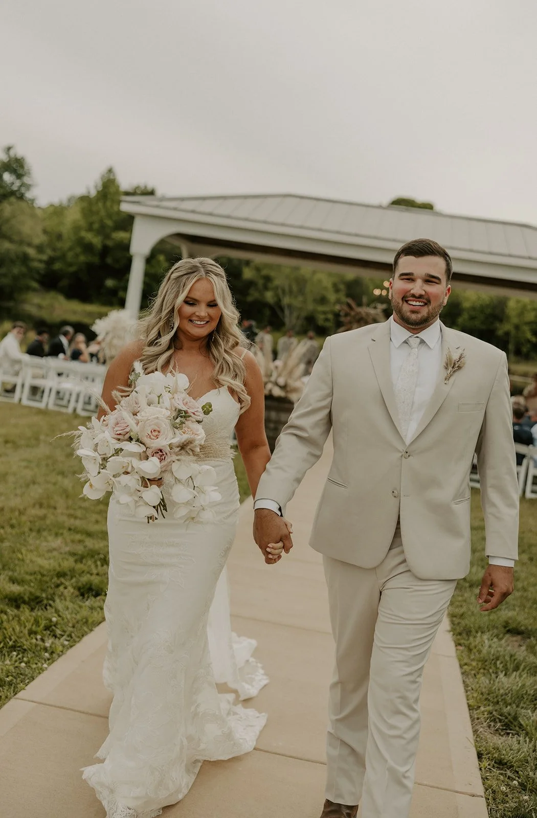A newlywed couple holding hands and walking outside during their wedding celebration. The bride is holding a large floral bouquet and is wearing a white lace wedding gown. The groom is wearing a light-colored suit and tie. There are guests seated on 