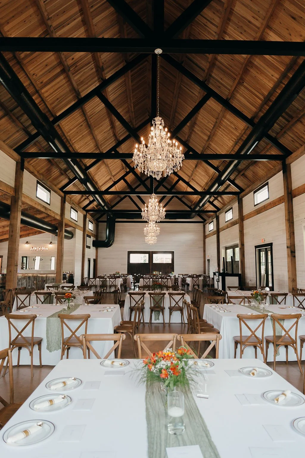 Interior of a rustic event venue with high wooden ceiling, chandeliers, and long tables with white tablecloths and floral centerpieces.