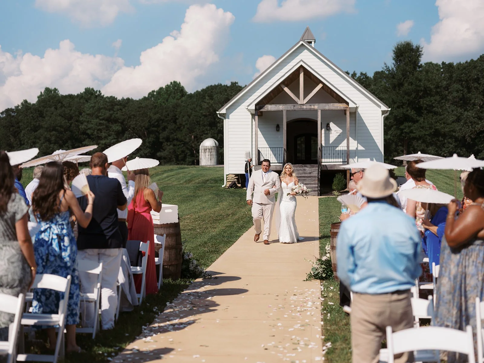 Bride walking down the aisle with her father at an outdoor wedding ceremony with guests seated on either side and holding parasols.