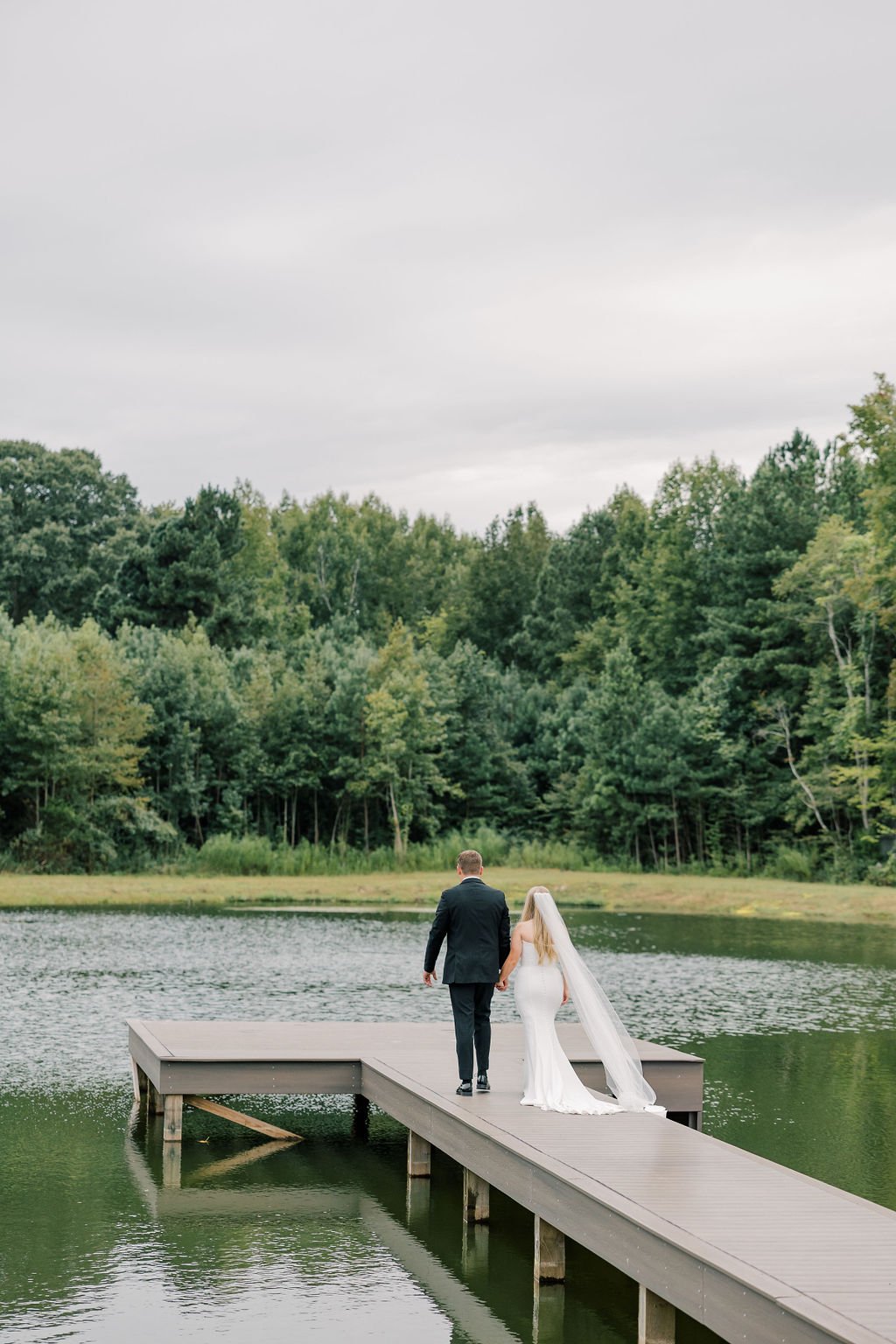 A bride and groom walking hand in hand on a wooden dock over a lake, with a backdrop of green trees and overcast sky.