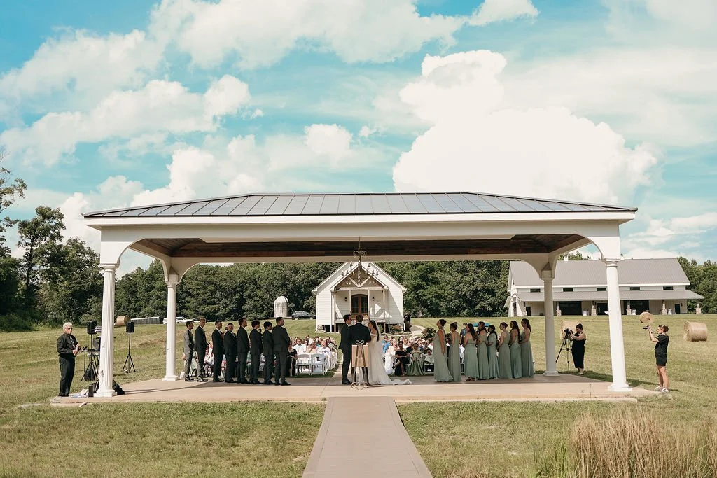 An outdoor wedding ceremony taking place under a pavilion with a rural farm backdrop, including a barn and hay bales, with guests, a bridal party, and photographers present.