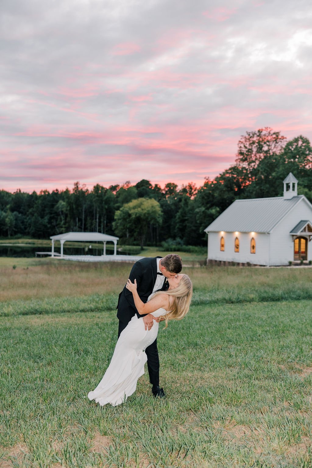A newlywed couple shares a romantic kiss outside during sunset, with a small white chapel and a pond in the background.