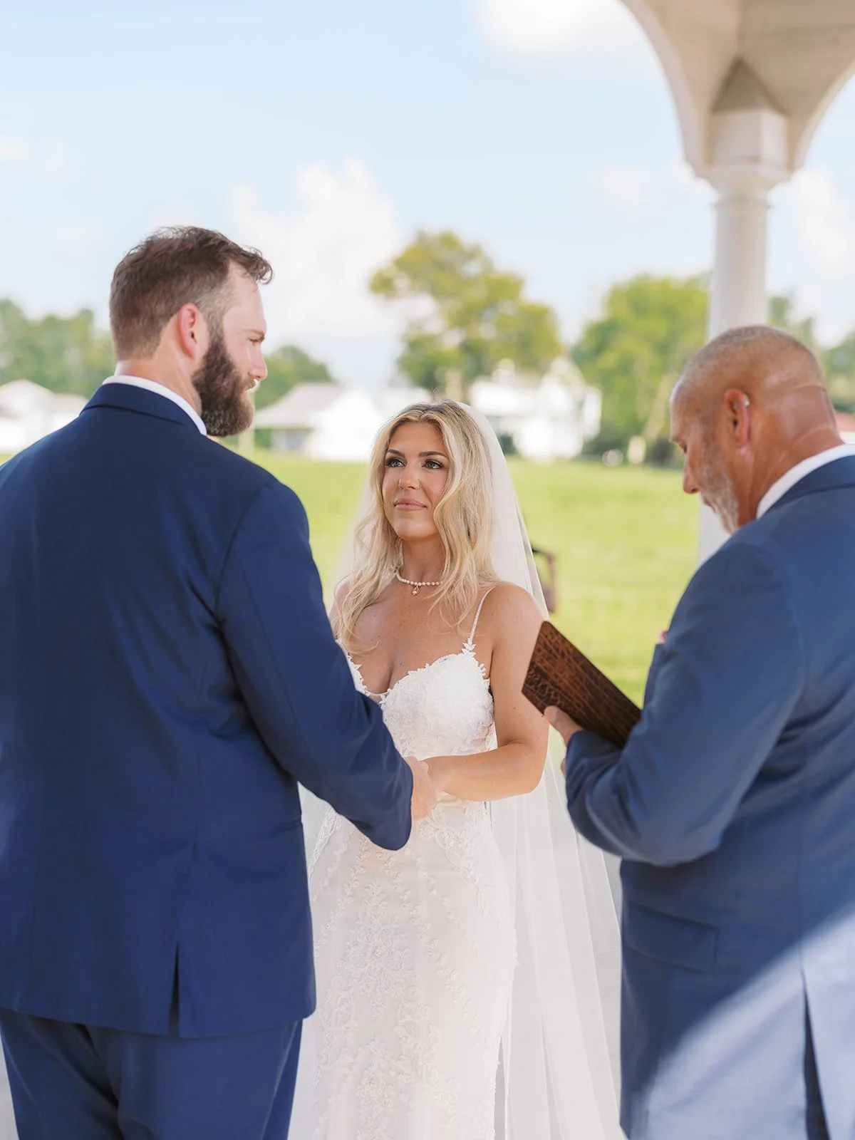 A wedding ceremony outdoors with a bride and groom holding hands, facing each other, while officiant reads from a book. The bride wears a white lace dress and veil, and the groom wears a navy suit. Trees and white buildings are in the background.