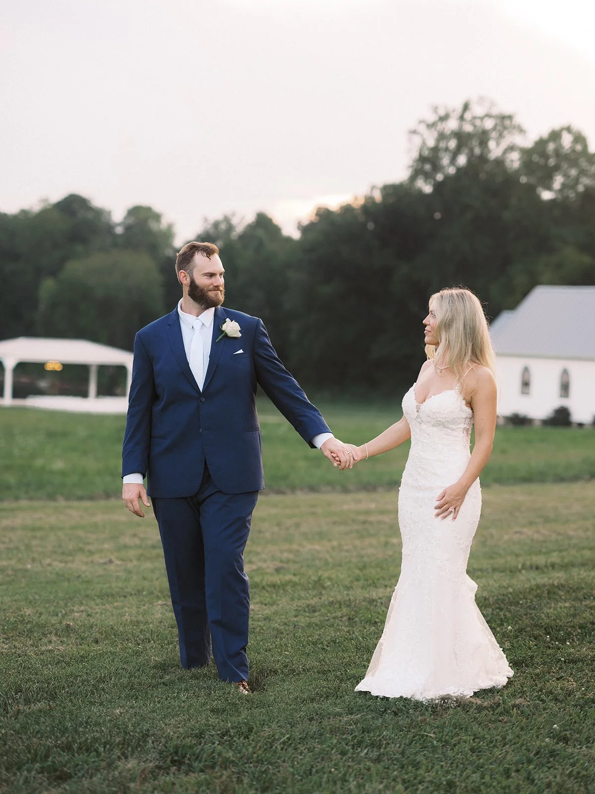 A bride and groom holding hands outdoors during a wedding, with trees and a white building in the background.