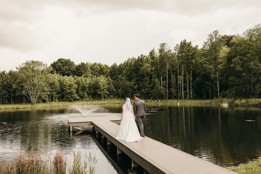 A bride and groom walk hand in hand on a wooden dock over a lake, with trees in the background, during a wedding photoshoot.