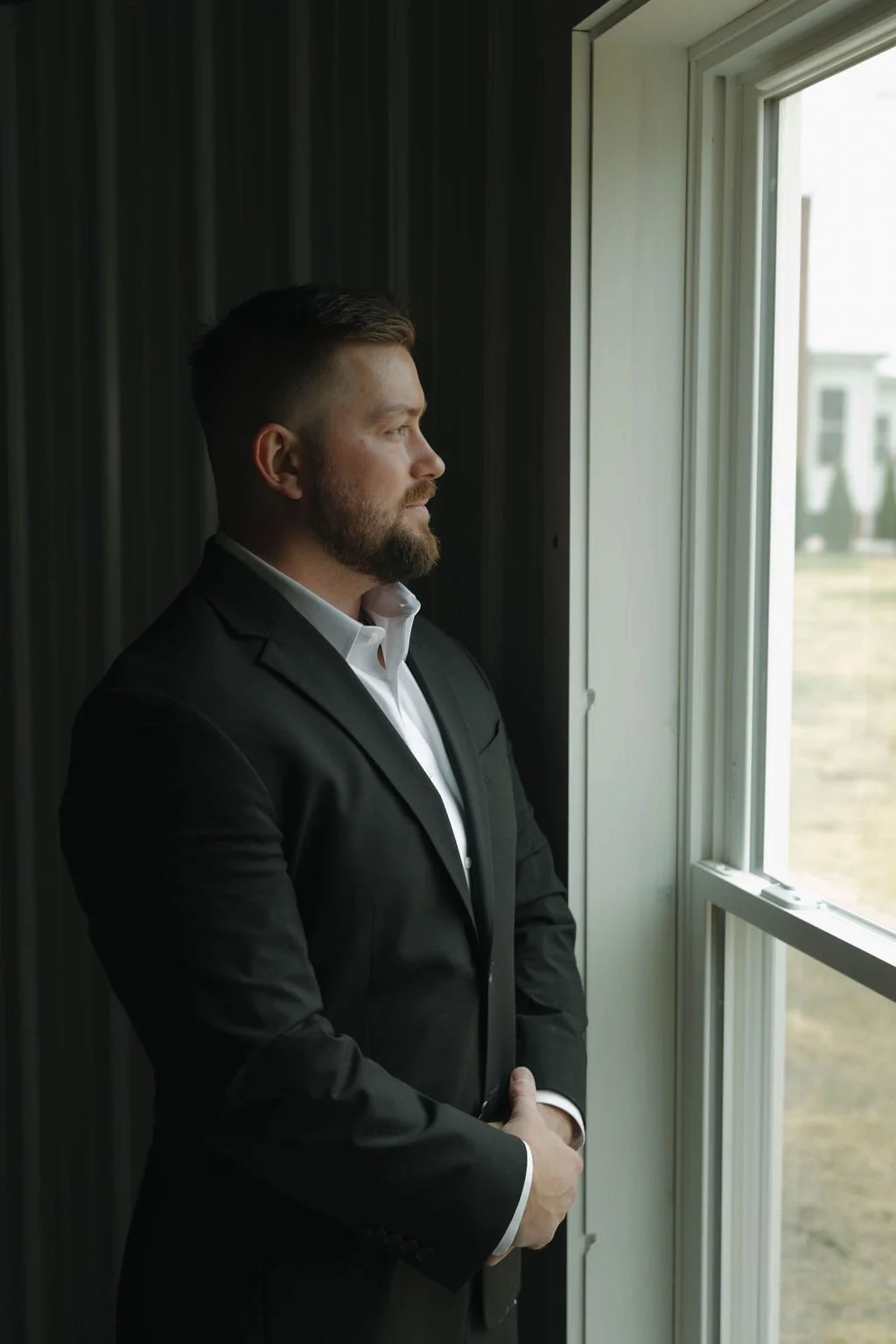 A man in a black suit and white shirt gazes out of a window, standing indoors with his arms crossed.