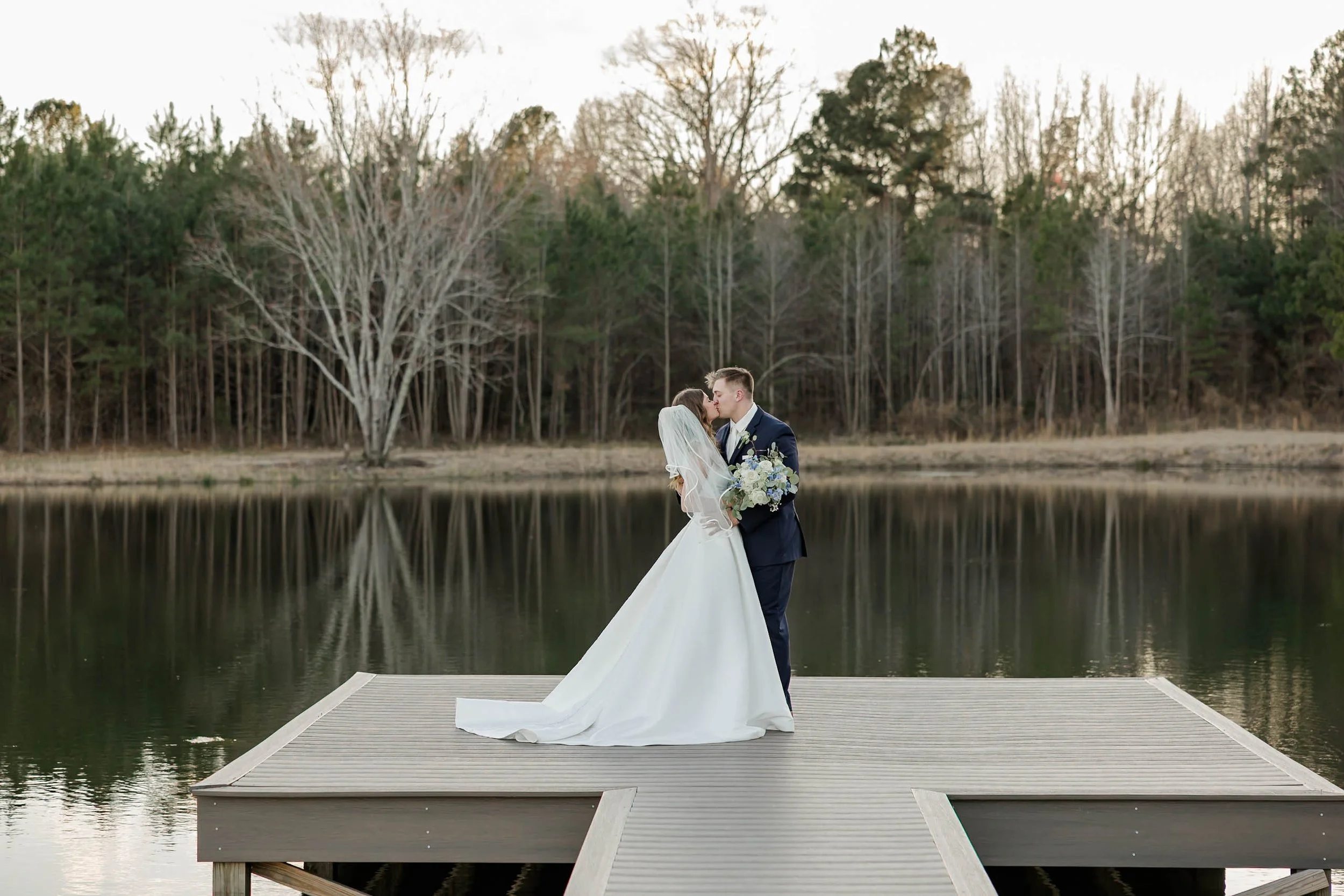 A bride and groom sharing a kiss on a dock by a lake with trees in the background.
