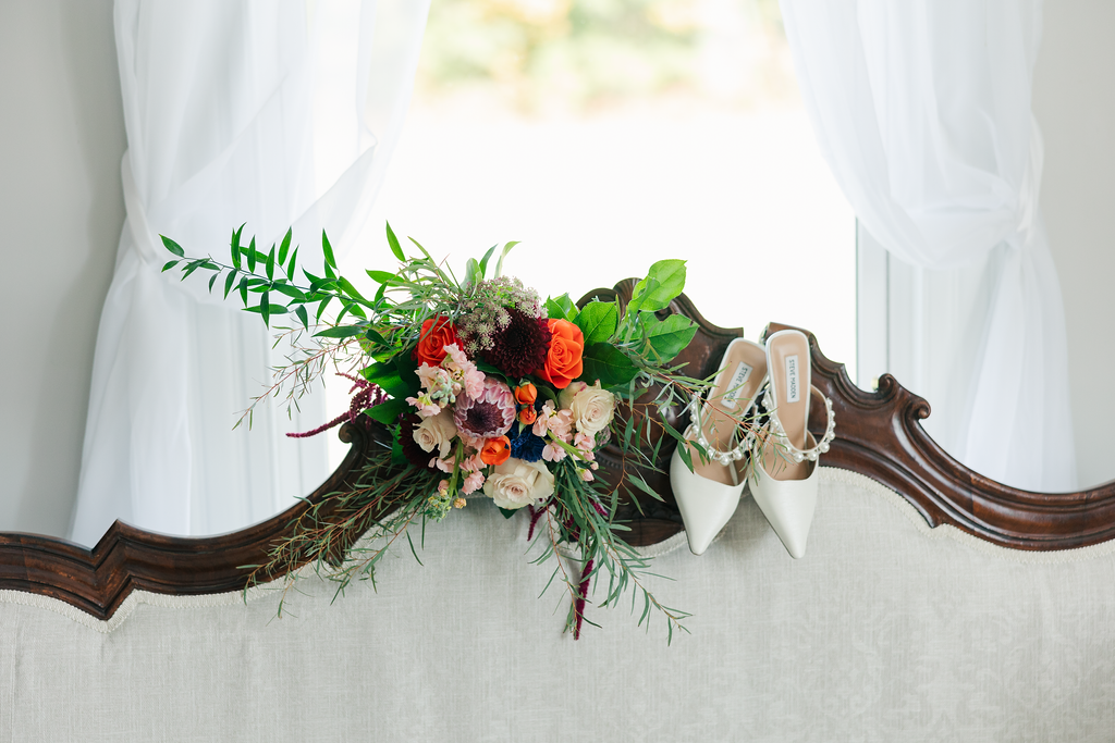 A bouquet of colorful flowers and a pair of white high heels resting on the back of a beige upholstered sofa near a window with white curtains.