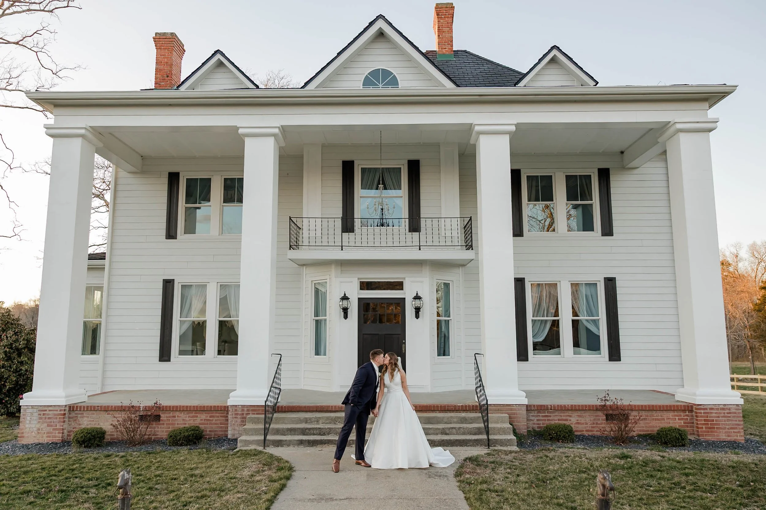 A newlywed couple kissing in front of a large white house with four tall pillars, a staircase, and multiple windows with black shutters. The bride is in a white wedding gown, and the groom is in a navy suit.