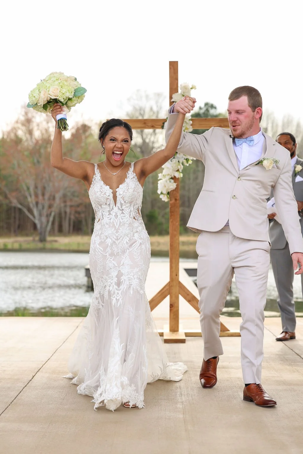 A newlywed couple celebrating their wedding outdoors by a lake, with the bride holding a bouquet and the groom raising her hand in victory. The bride is wearing a lace wedding dress and the groom a light-colored suit with a bowtie.
