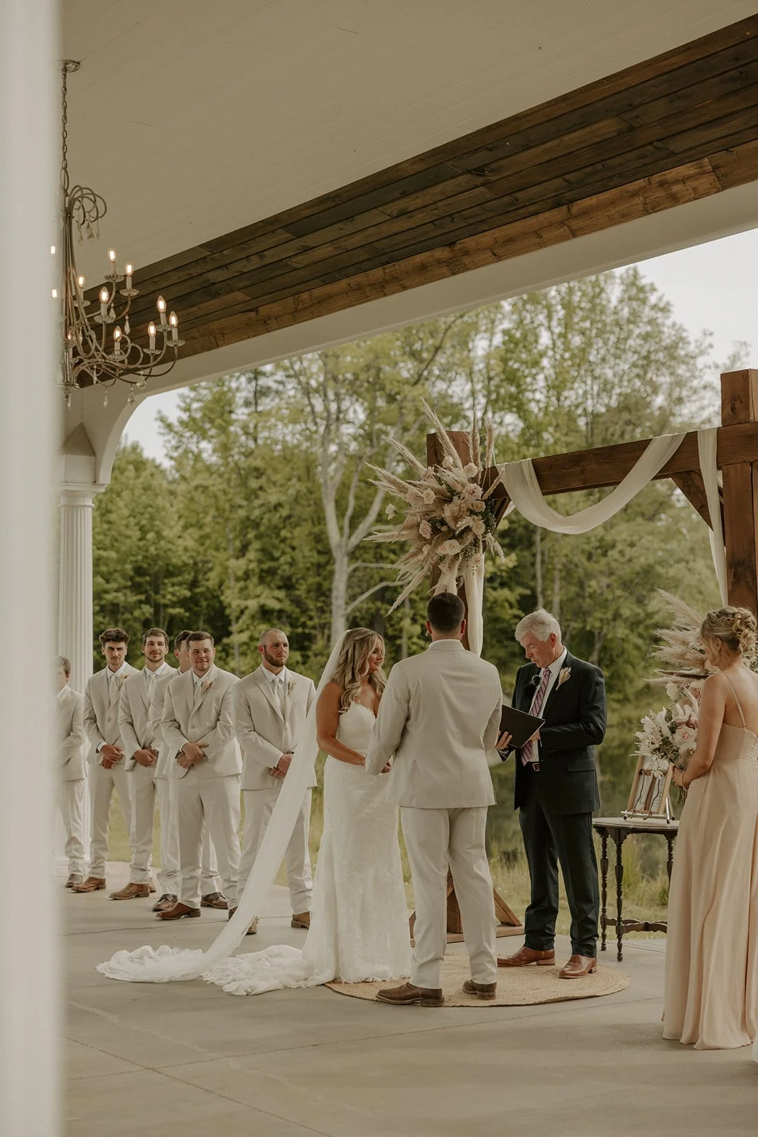A wedding ceremony taking place outdoors under a wooden arch decorated with white and pink flowers and draped fabric, with the bride and groom exchanging vows in front of an officiant, and a bridesmaid holding a bouquet on the right side.