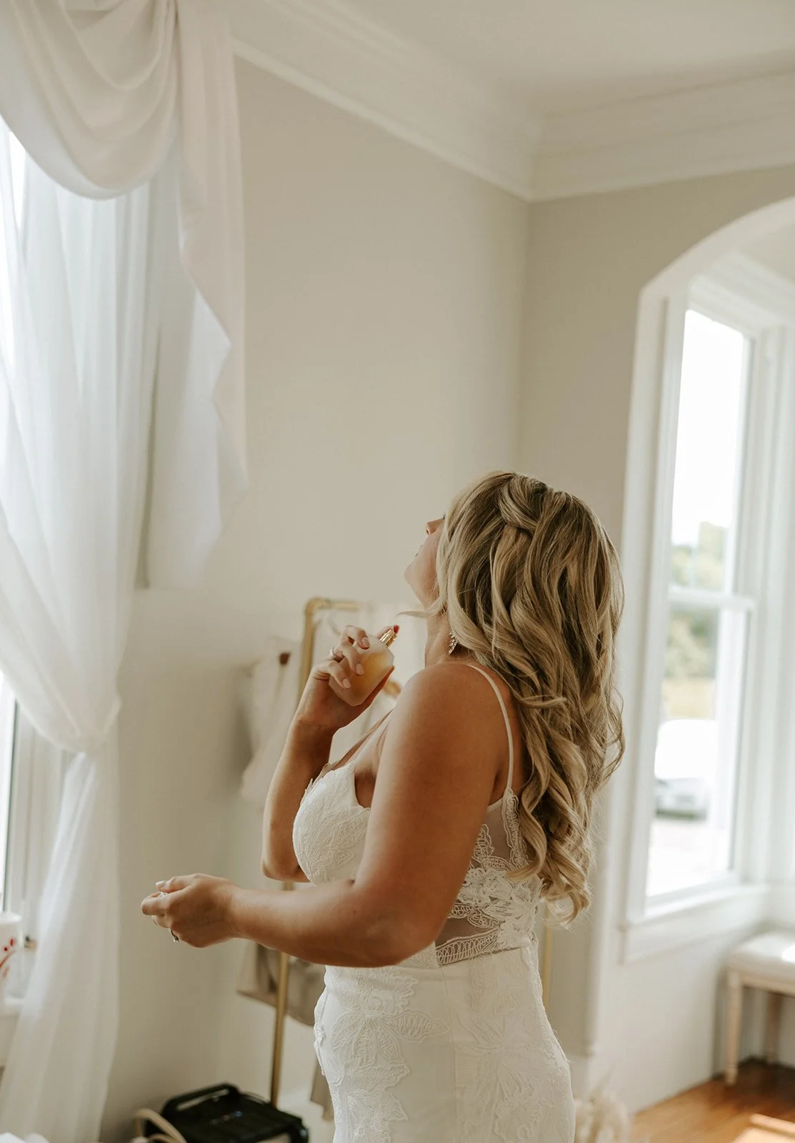 A woman with long blonde hair in a white lace wedding dress is spraying perfume near her neck in a bright room with large windows and white curtains.