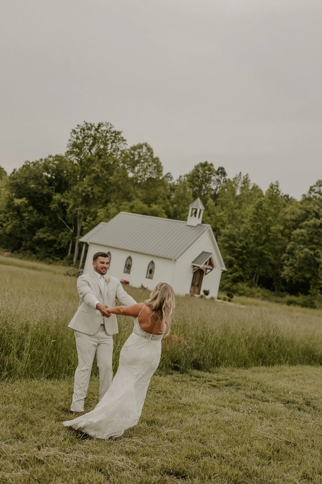 A couple in wedding attire smiling and holding hands, dancing outdoors in front of a small white chapel surrounded by green trees and grass.