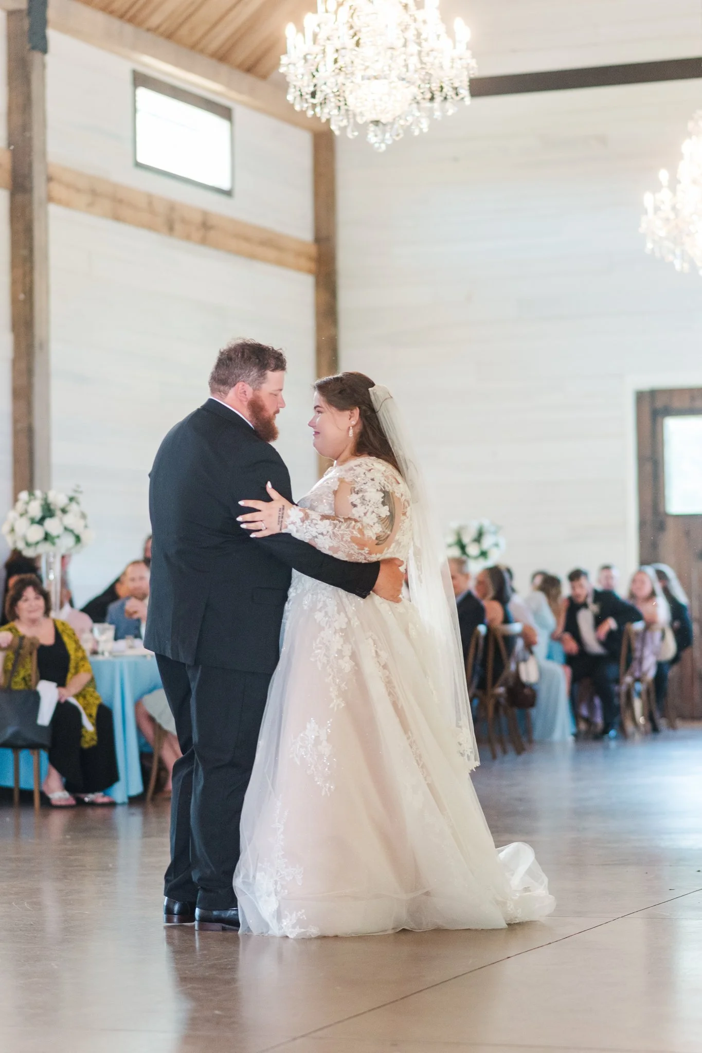 A bride and groom sharing their first dance at a wedding reception, with guests in the background and chandeliers hanging from the ceiling.