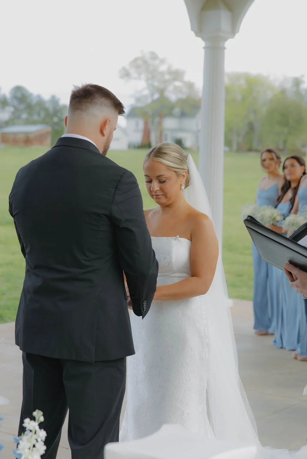 Bride and groom exchanging vows at an outdoor wedding ceremony under a white pergola, with bridesmaids in blue dresses in the background.