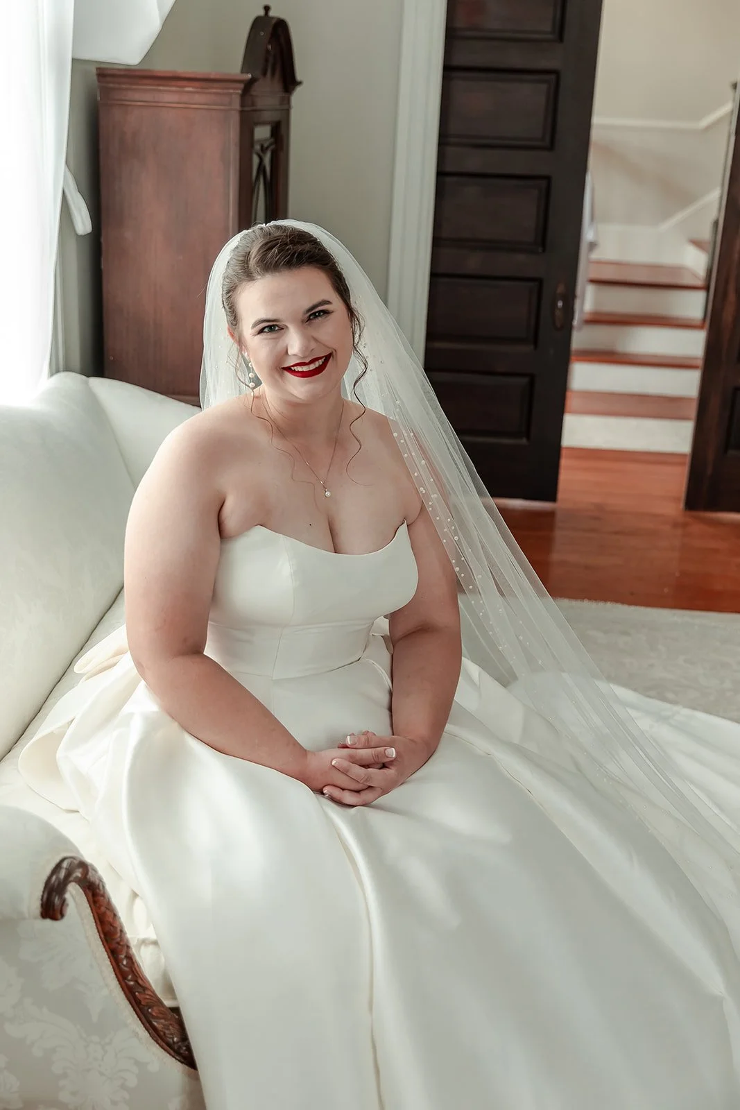 A smiling bride in a white wedding dress with a veil sitting on a cream-colored sofa in a well-lit room with wooden furniture and stairs in the background.