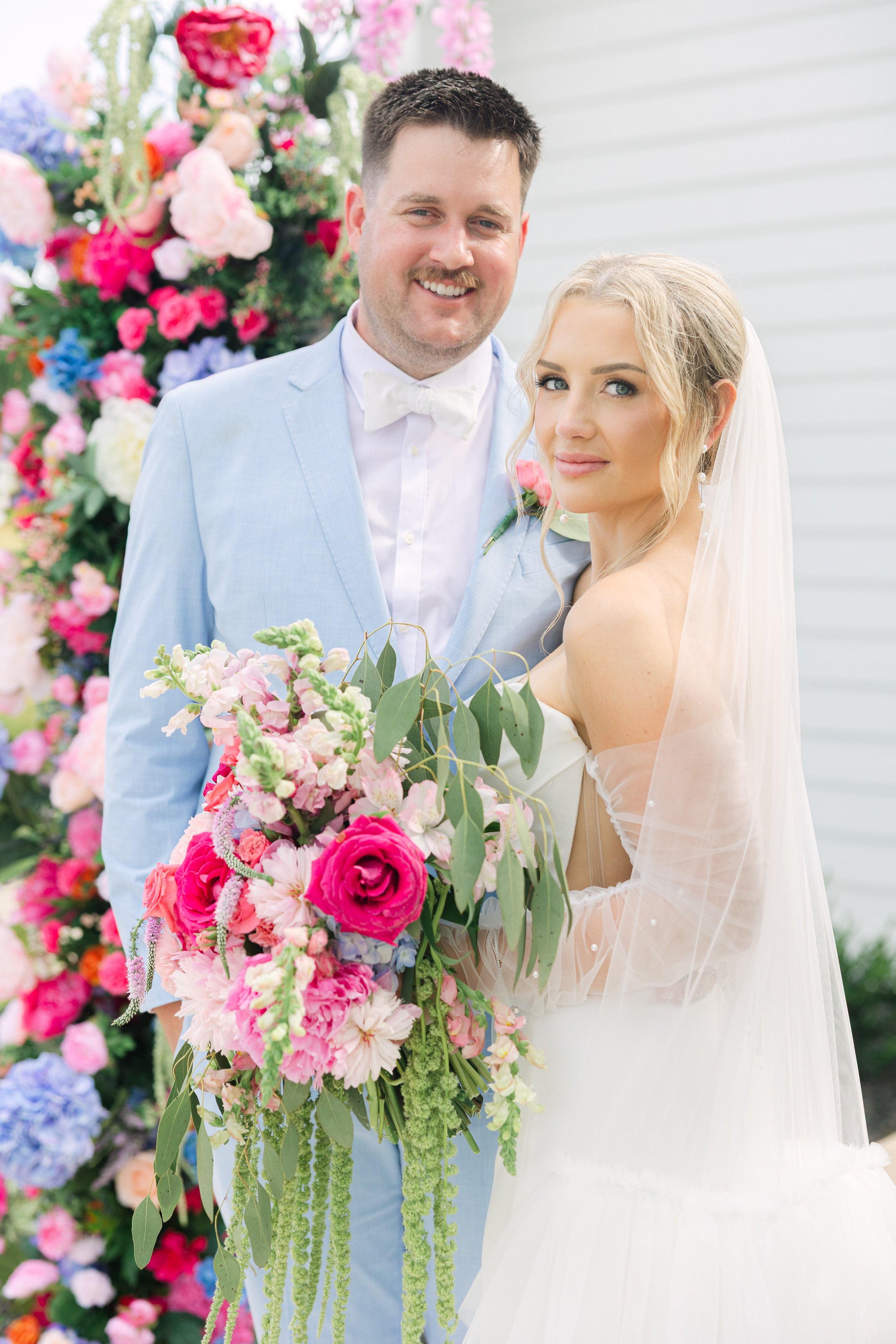 A newlywed couple standing outdoors, surrounded by colorful flowers. The groom is wearing a light blue suit with a white shirt and bow tie. The bride is wearing a white wedding dress with sheer, beaded long sleeves, and a veil. She is holding a large