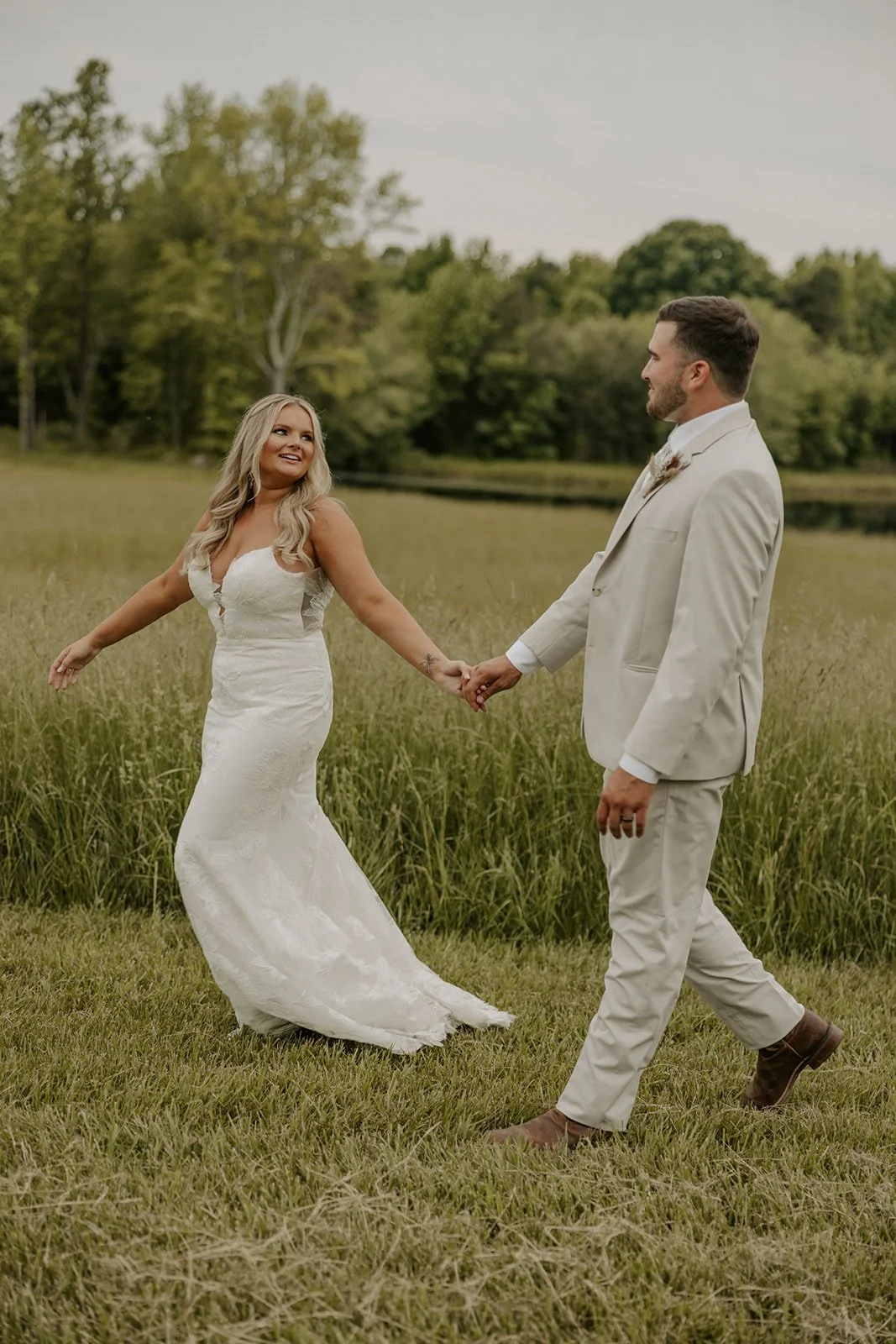 A couple in wedding attire holding hands and dancing outdoors on a grassy field near a pond with trees in the background.