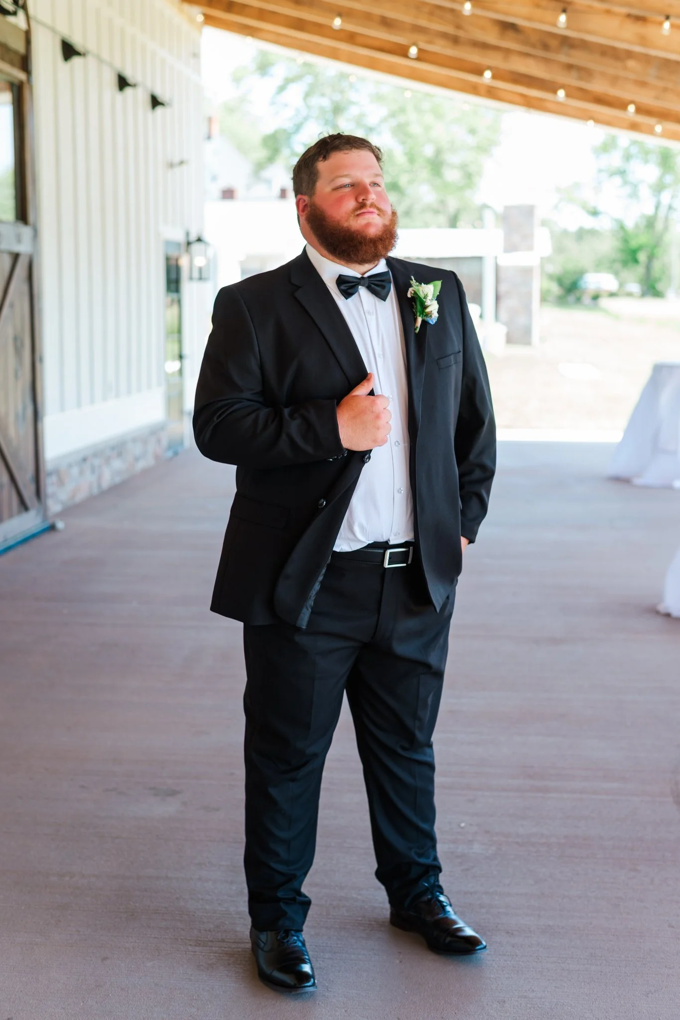 Groom in a black tuxedo with a bow tie and boutonniere, standing confidently with his hand on his chest in a wedding venue.