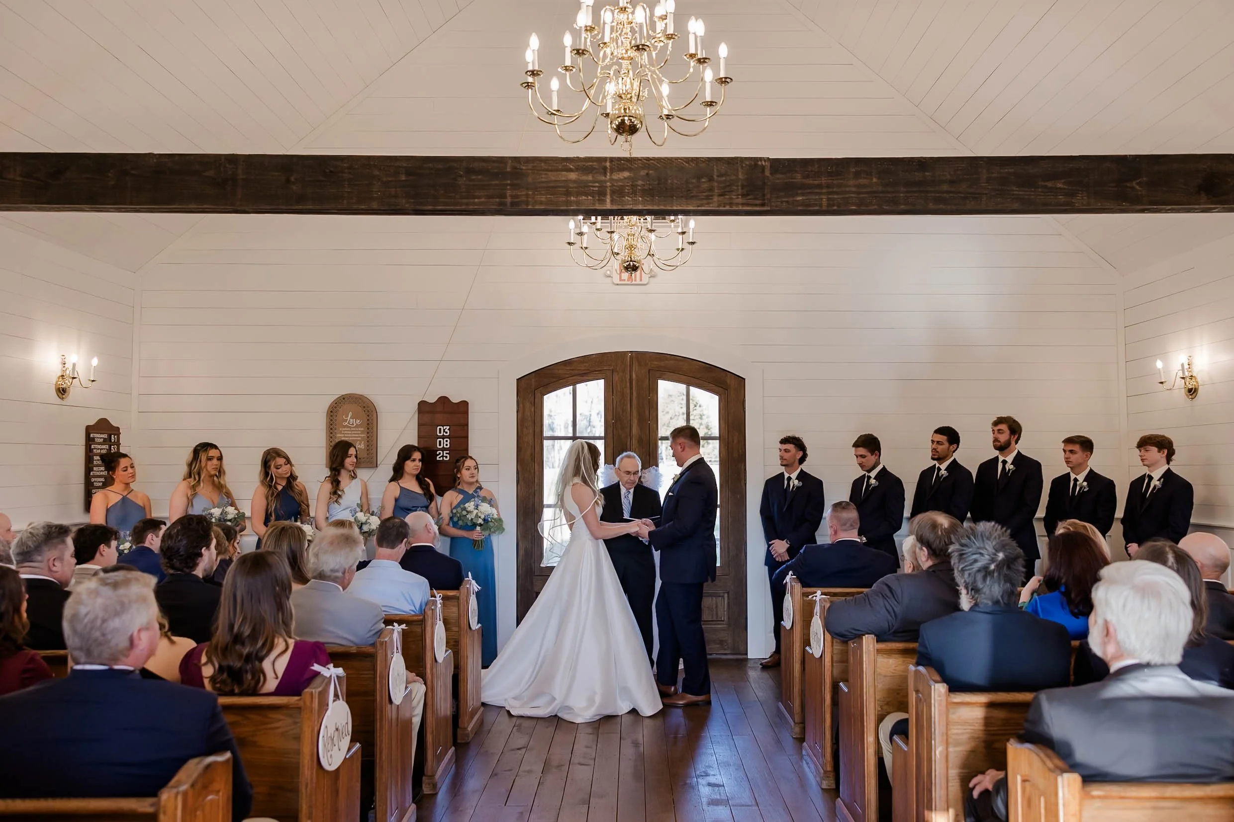 A wedding ceremony inside a church with a bride and groom facing each other and holding hands, surrounded by attendants and guests. The interior has white walls, wooden accents, chandeliers, and wall-mounted light fixtures.
