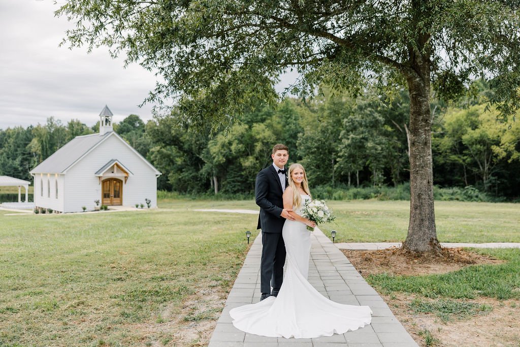 A bride and groom standing on a paved walkway in front of a tree, with a small white chapel in the background, during a wedding outdoors.