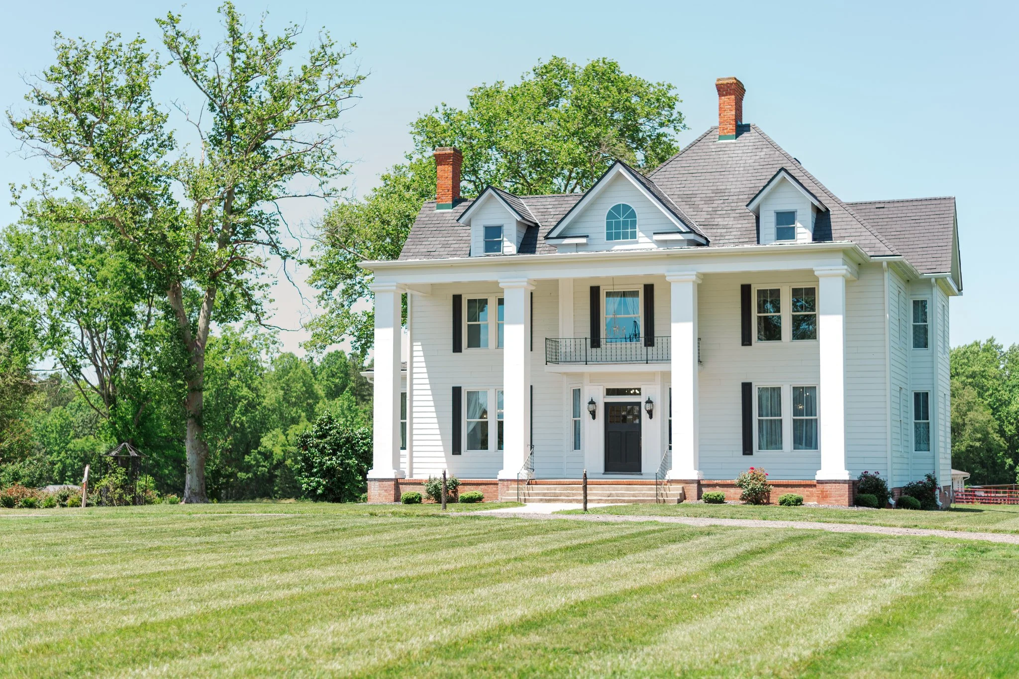 Large white house with columns on the front porch and black shutters, surrounded by green trees and grass under a clear blue sky.