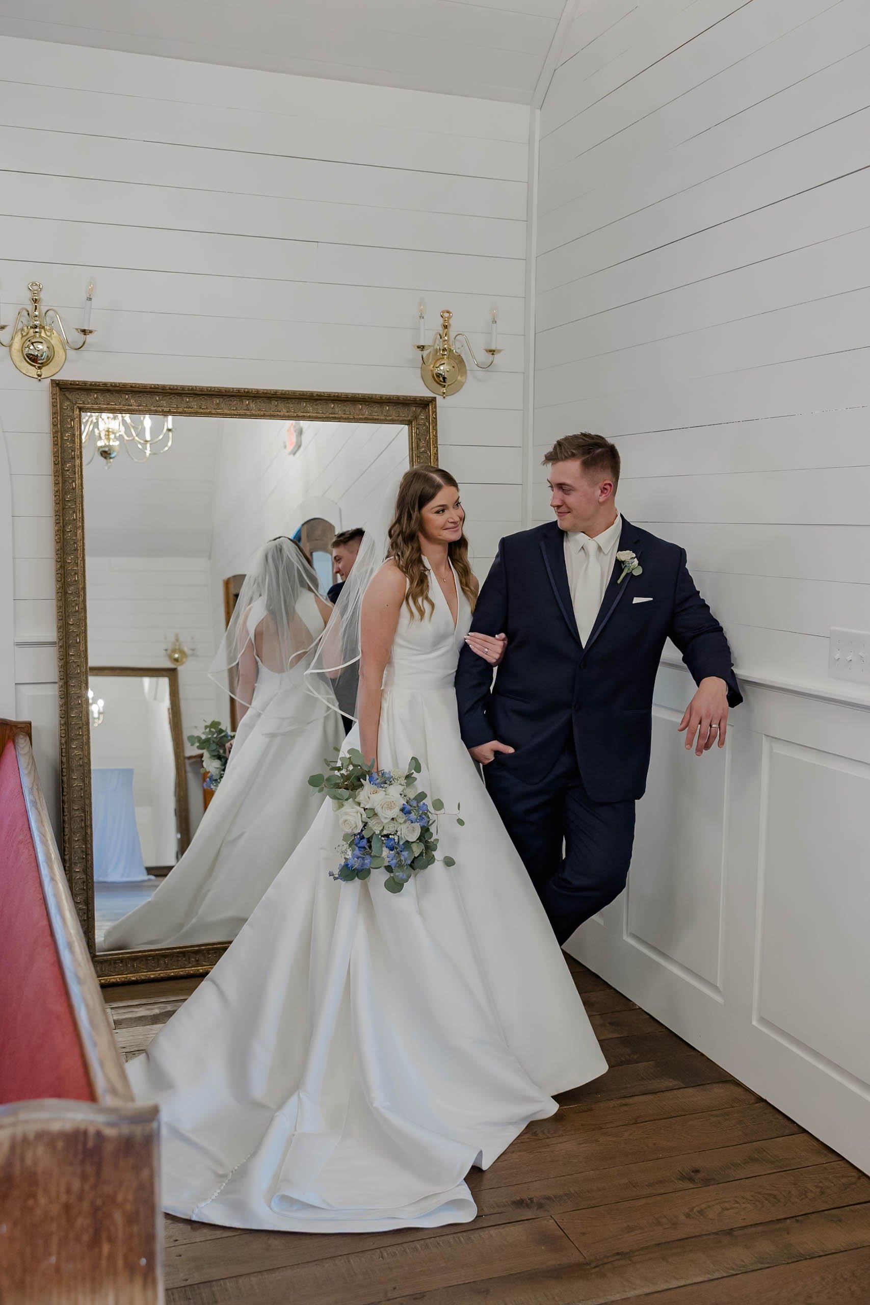 Bride in a white wedding dress holding a bouquet standing next to groom in a dark suit inside a room with white paneled walls, gold sconces, and a large mirror reflecting both of them.