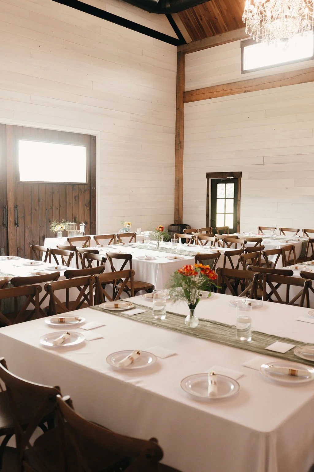A rustic event space with wooden walls and ceiling, set up for a gathering with long tables covered in white tablecloths, decorated with small flower arrangements and lined with wooden chairs.