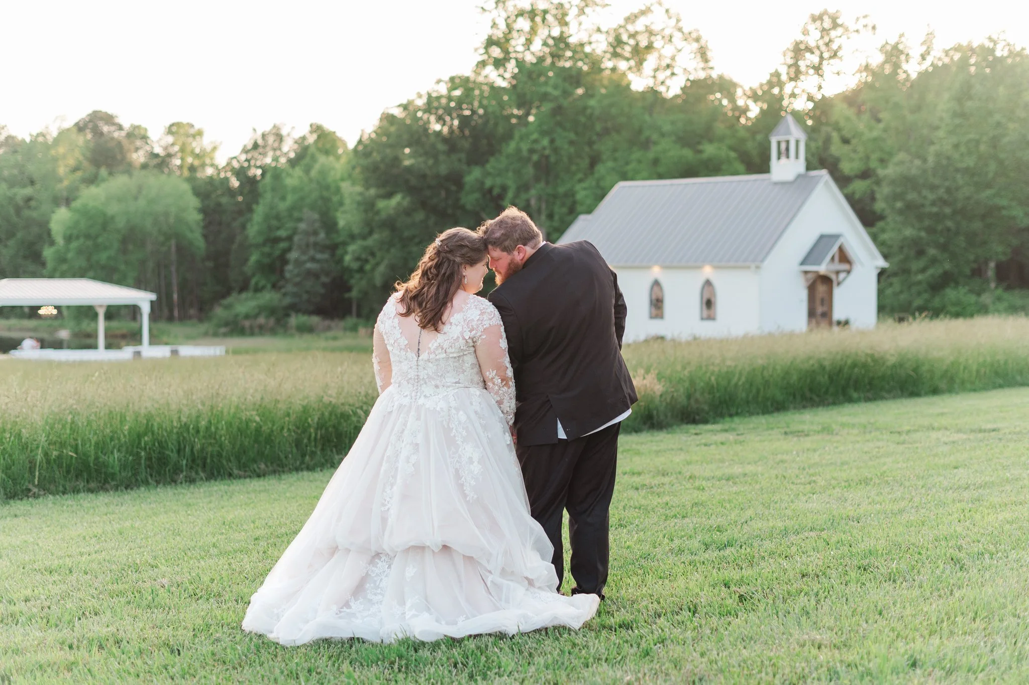 A bride and groom standing closely on a grassy field near a small white chapel, sharing an intimate moment with their foreheads touching, during their wedding day at sunset.