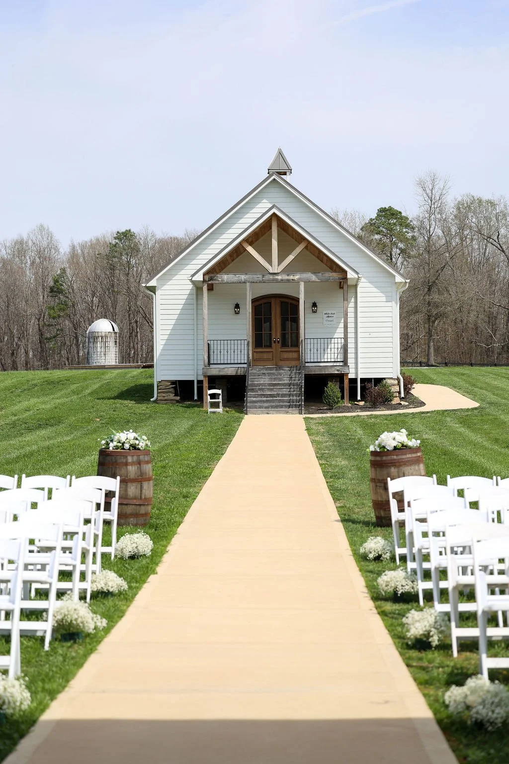 Small white chapel with wooden front door and stairs, set outdoors on a grassy lawn, decorated for a wedding with white chairs and flowers along the aisle.