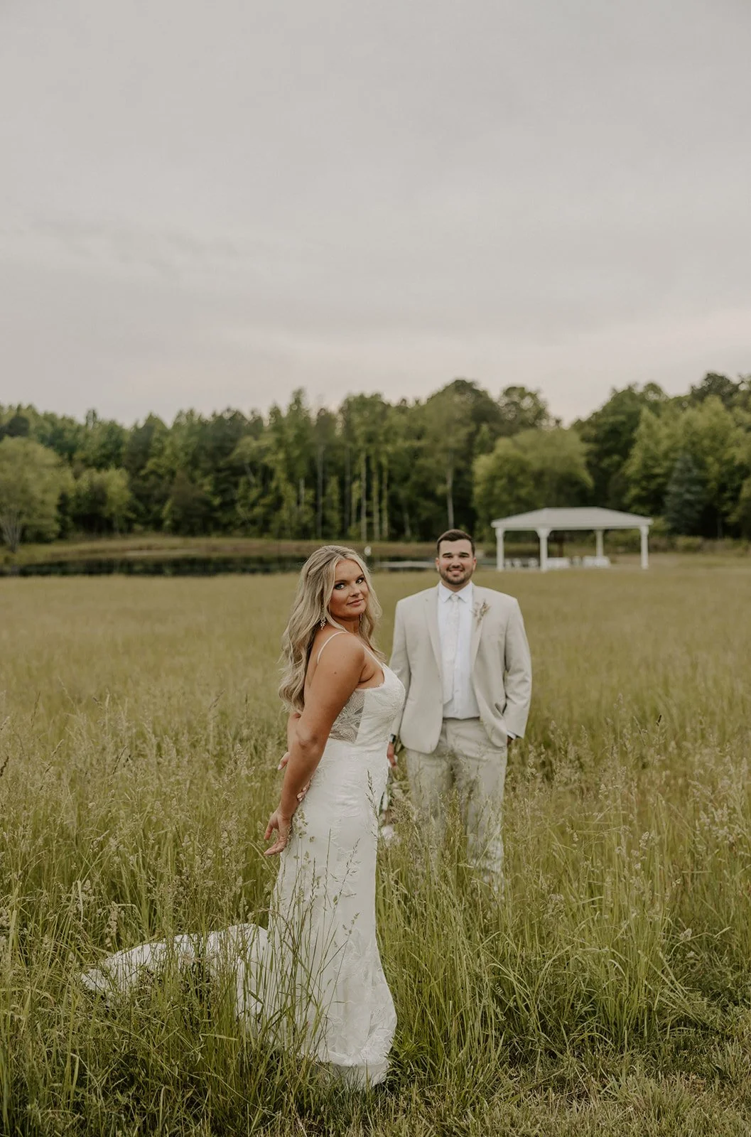 A bride in a white lace wedding gown and a groom in a light-colored suit standing in a grassy field with trees and a white pavilion in the background.