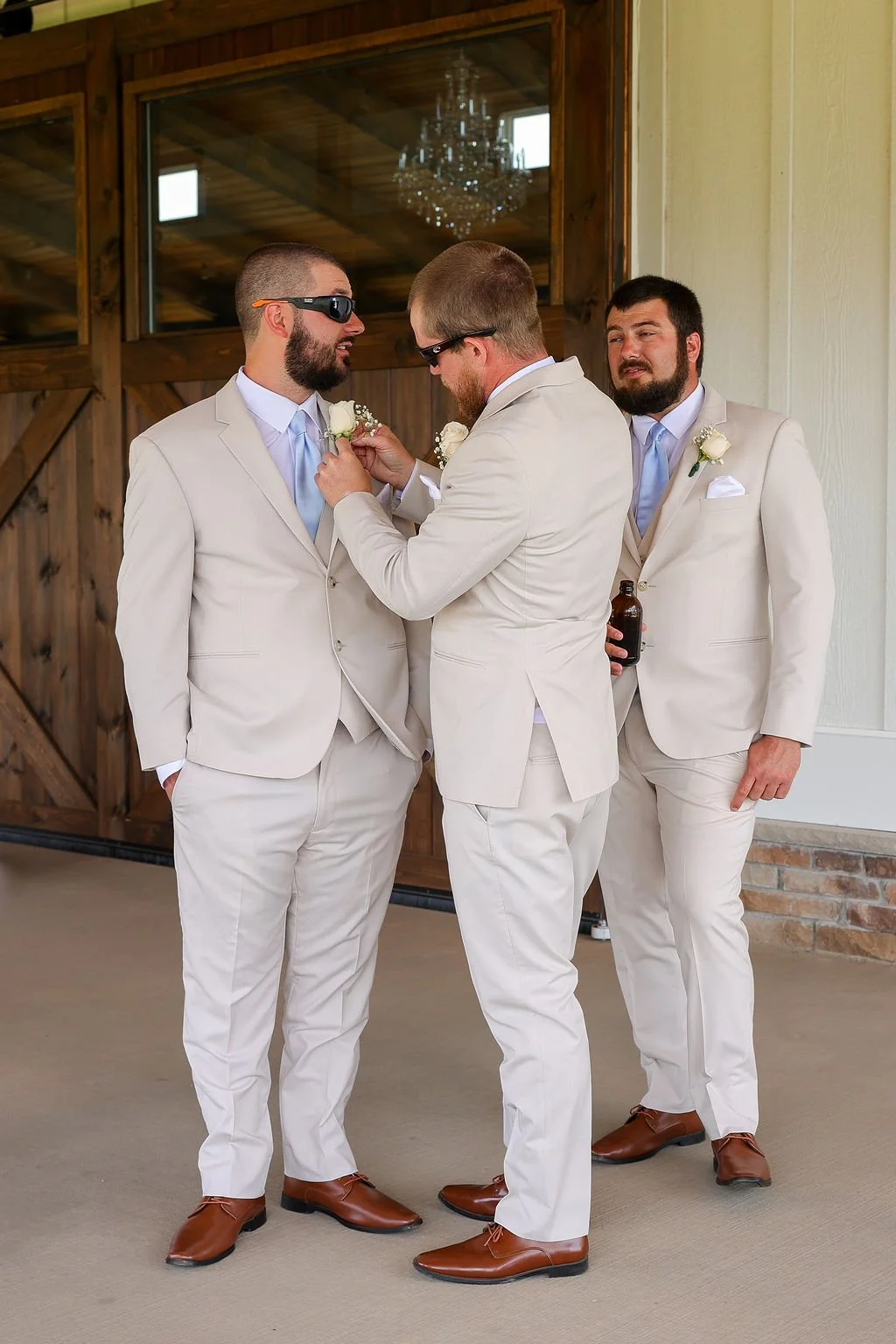 Three men dressed in beige suits and brown shoes, with two wearing sunglasses and boutonnieres, standing indoors in front of wooden sliding doors. One man is pinning a boutonniere onto another.