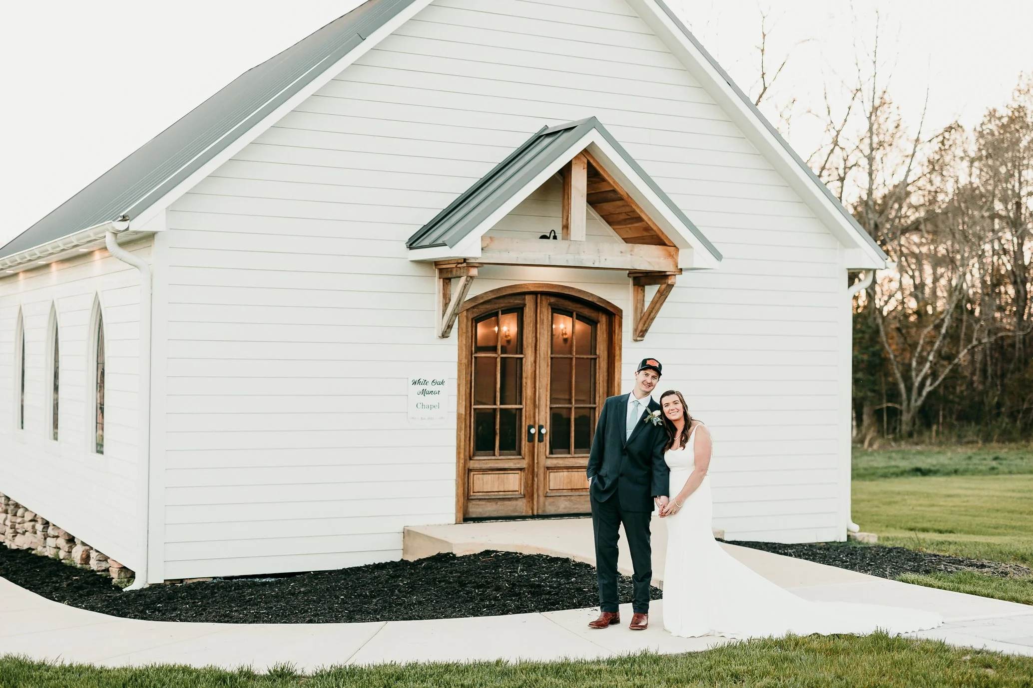 A bride and groom standing outside a small white chapel, holding hands and smiling, with the bride wearing a white wedding dress and the groom in a dark suit, in a grassy area during sunset.