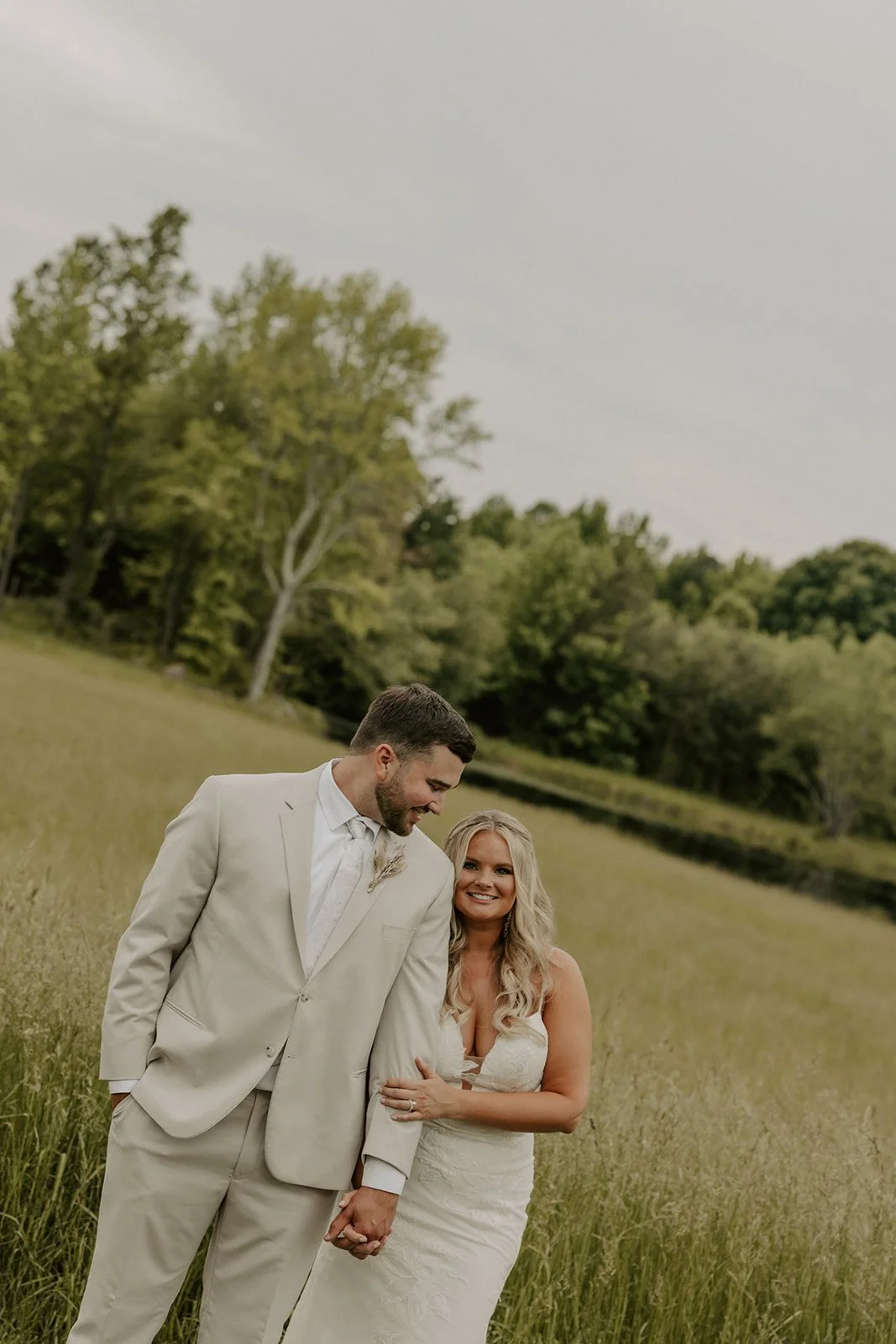 A newlywed couple holding hands in a grassy field with trees in the background. The groom is wearing a light beige suit and the bride is in a white lace wedding dress, smiling at each other.