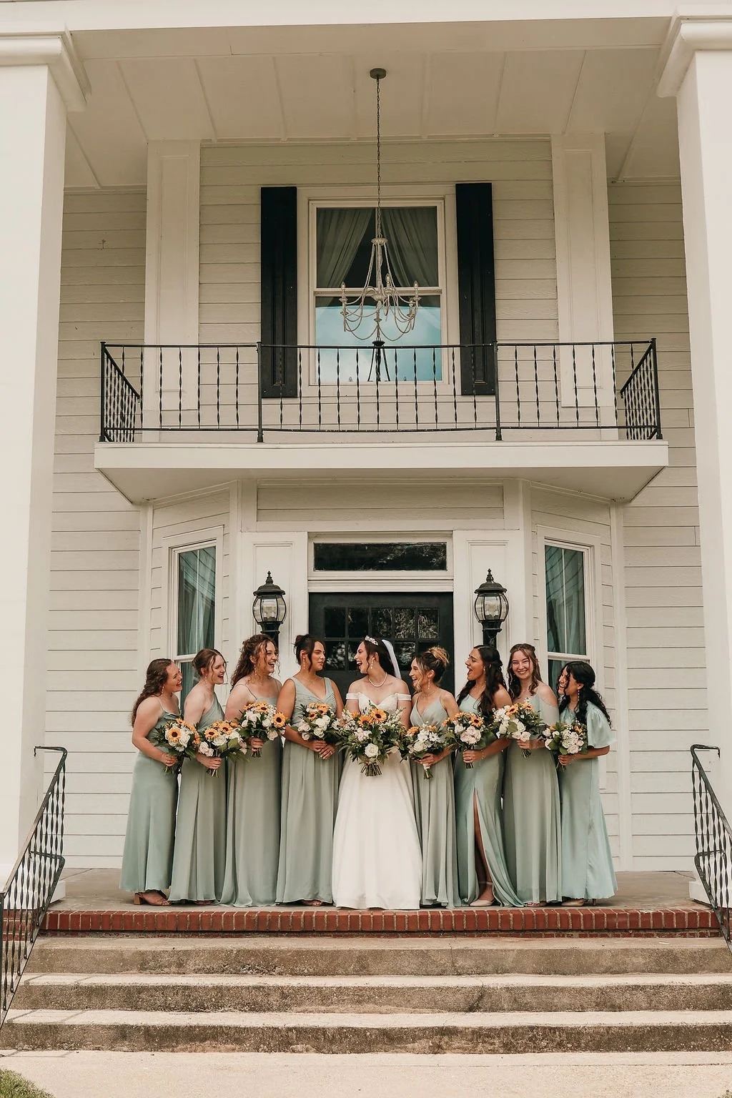 A bride and eight bridesmaids standing on steps in front of a white house with black lanterns. The women are holding bouquets, and the bride is wearing a white dress with an off-the-shoulder neckline and tiara.