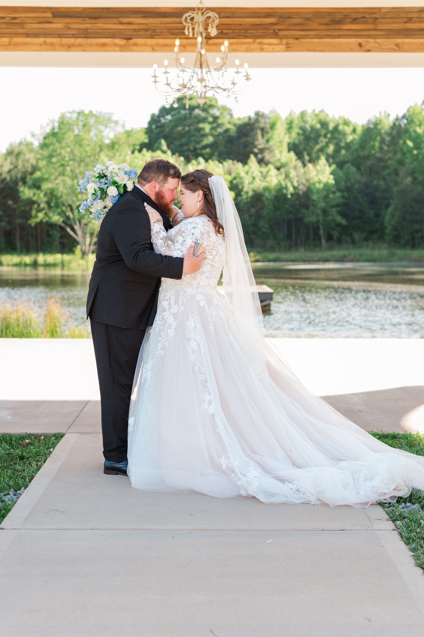 A bride and groom on their wedding day, standing close with foreheads touching, outdoors near a lake, under a wooden pavilion with a chandelier, greenery in the background.