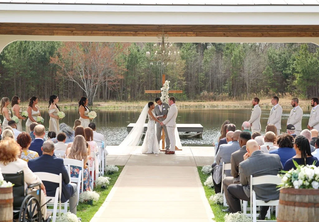 A wedding ceremony takes place outdoors next to a lake, with the bride and groom exchanging vows under a wooden cross decorated with flowers, surrounded by bridesmaids and groomsmen, and seated guests in front.