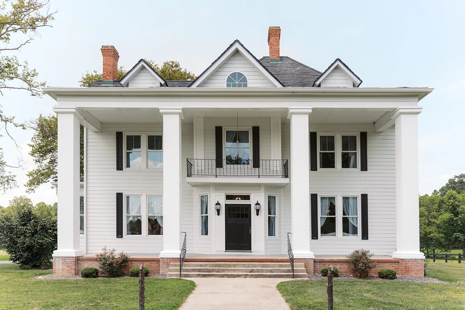 White two-story house with black shutters, brick foundation, and a black front door, surrounded by a green lawn and trees.