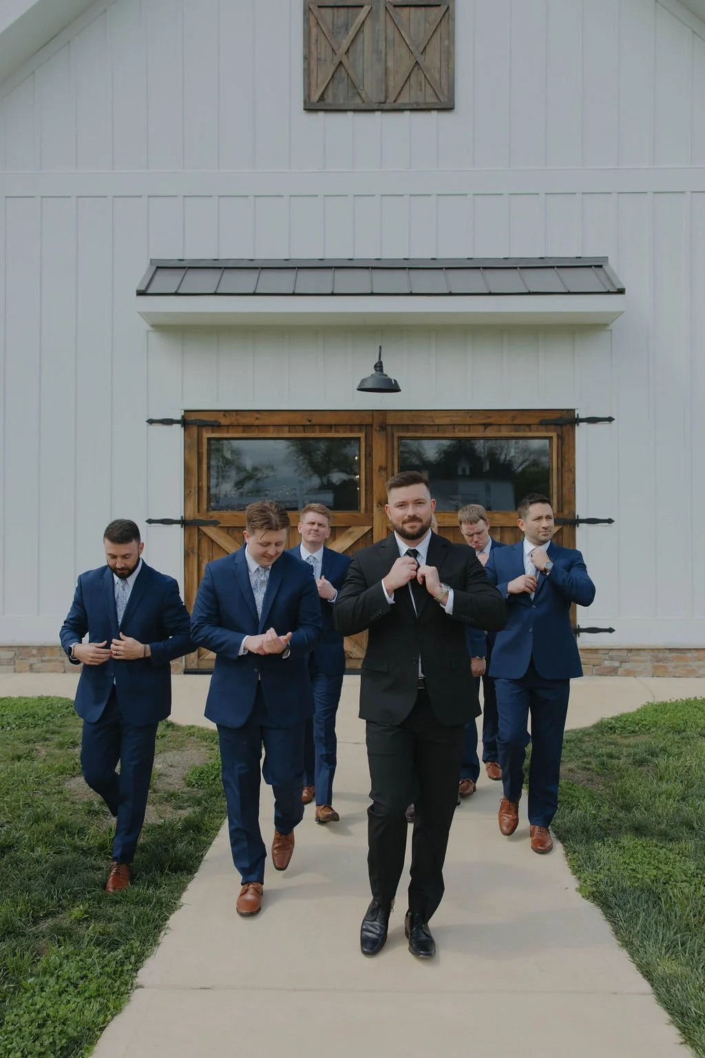 A group of six men in suits walking in front of a white barn with wooden doors and black metal accents, with the groom in a black suit leading and the groomsmen behind in blue suits.
