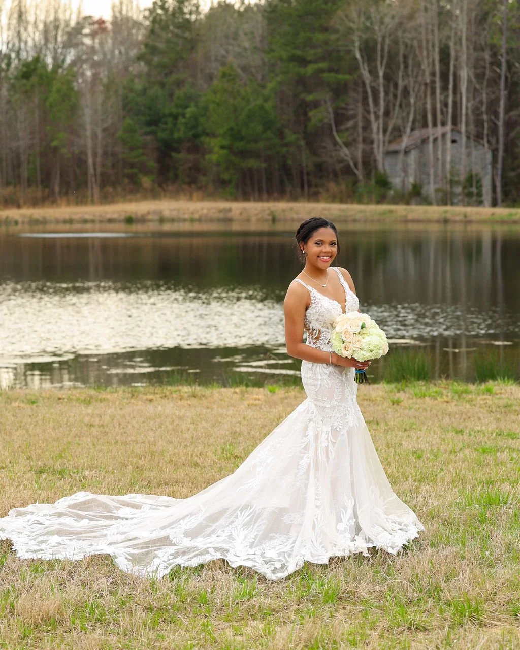 A smiling bride in a lace wedding dress standing on grass near a lake, holding a bouquet of white and pale pink roses, with trees and a small building in the background.