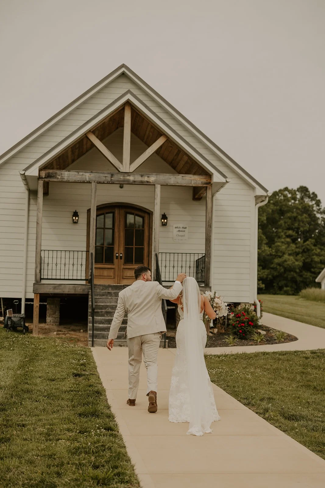A bride and groom walking towards a white church, with the groom touching the bride's veil, on a sidewalk with grass on both sides.