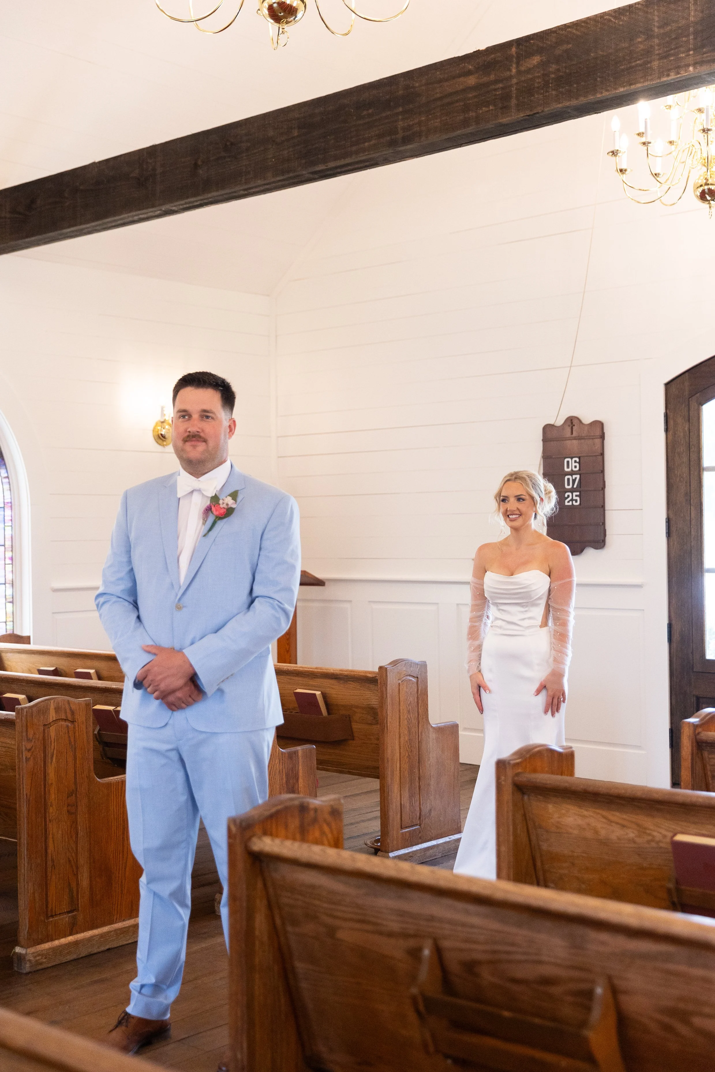 A groom in a light blue suit and a bride in a white wedding gown standing in a church with wooden pews, chandeliers, and a wall-mounted hymn board.