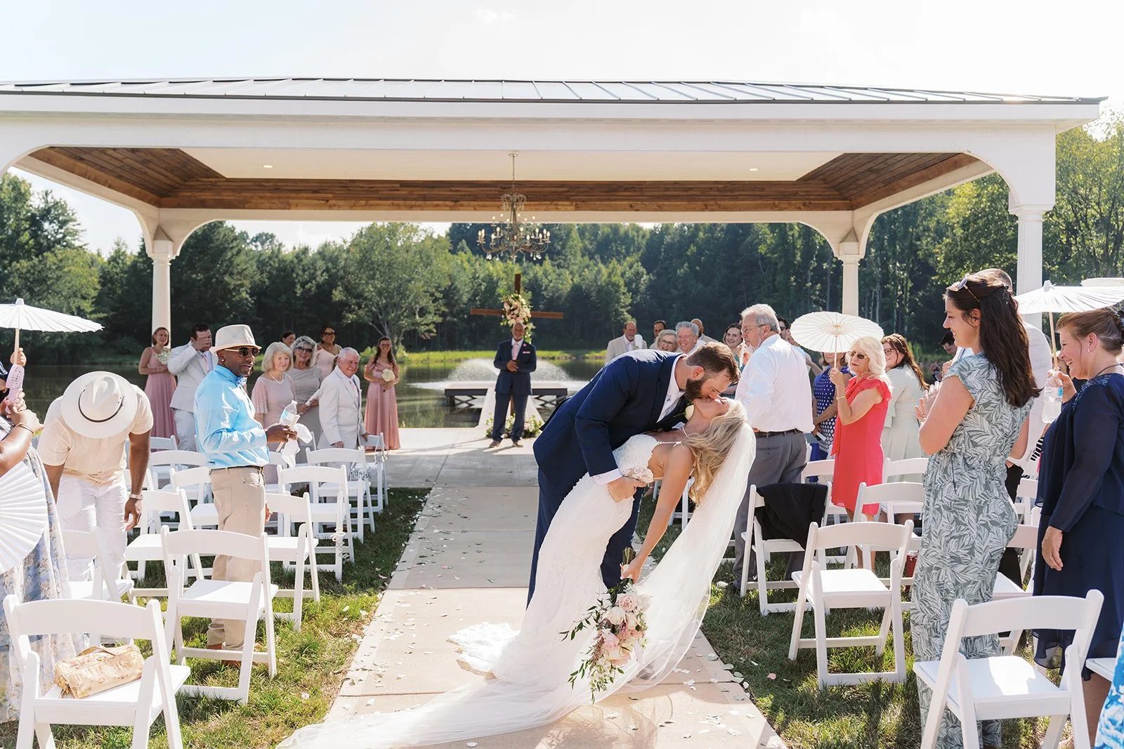 A newlywed couple sharing a kiss during an outdoor wedding ceremony, with guests sitting and standing around them holding umbrellas and celebrating under a pavilion near a lake.