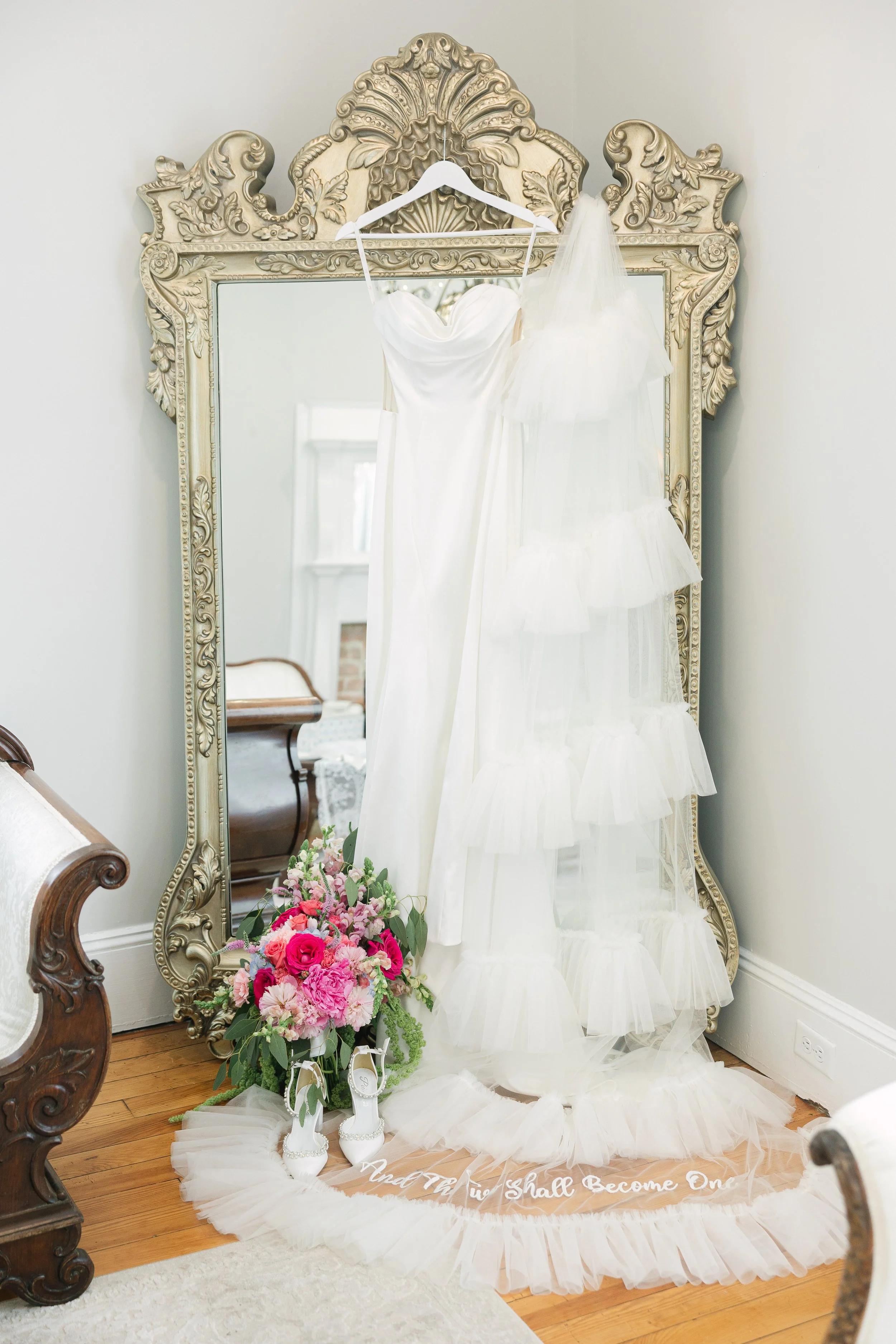 A white wedding dress hanging on a white hanger on an ornately framed mirror, with a veil draped over the dress. A bouquet of pink and purple flowers with white shoes placed in front, and a light-colored tulle fabric with white writing on the floor.