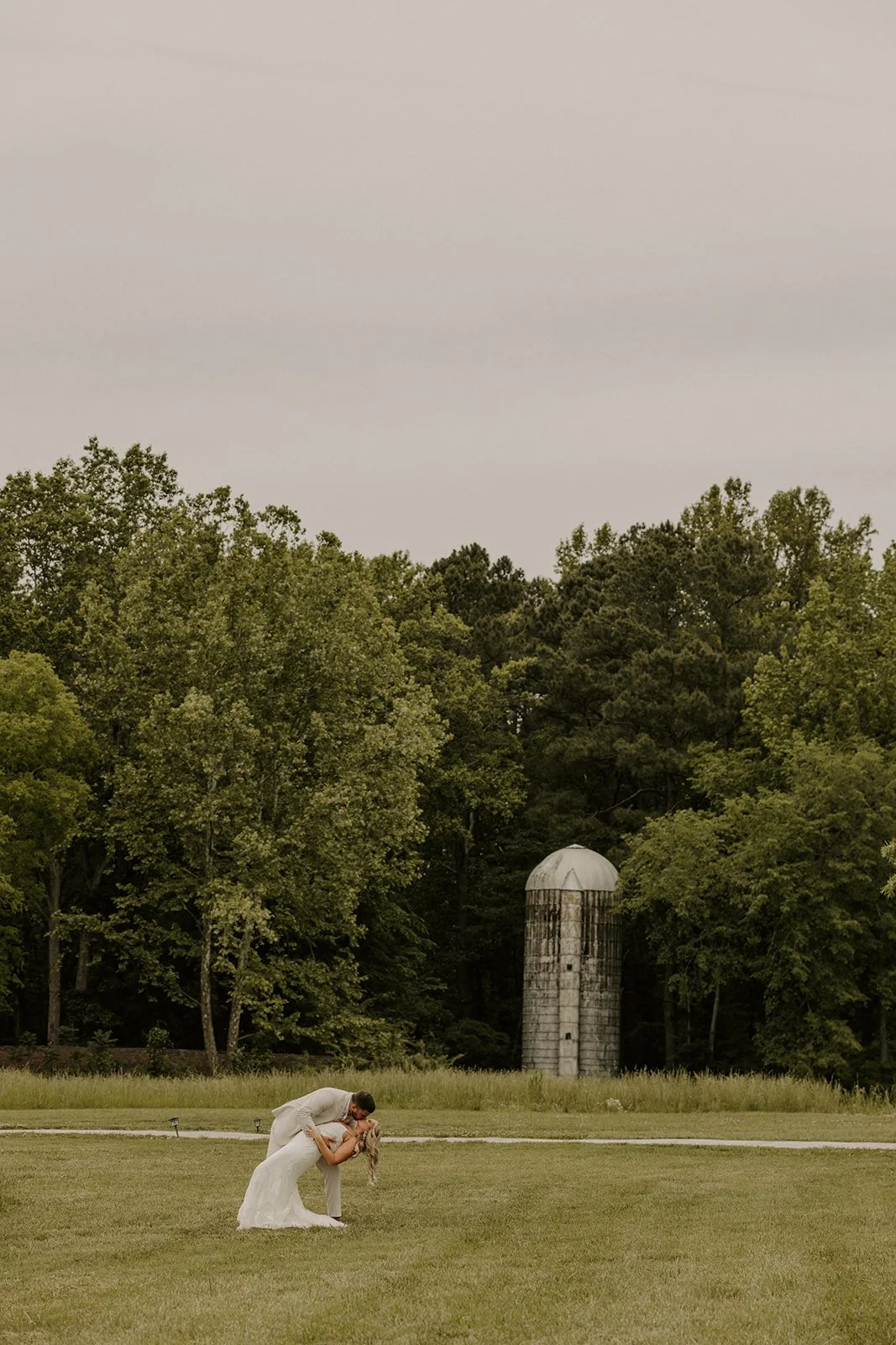 A bride and groom dancing outdoors on a grassy field with trees and a tall silo in the background on an overcast day.