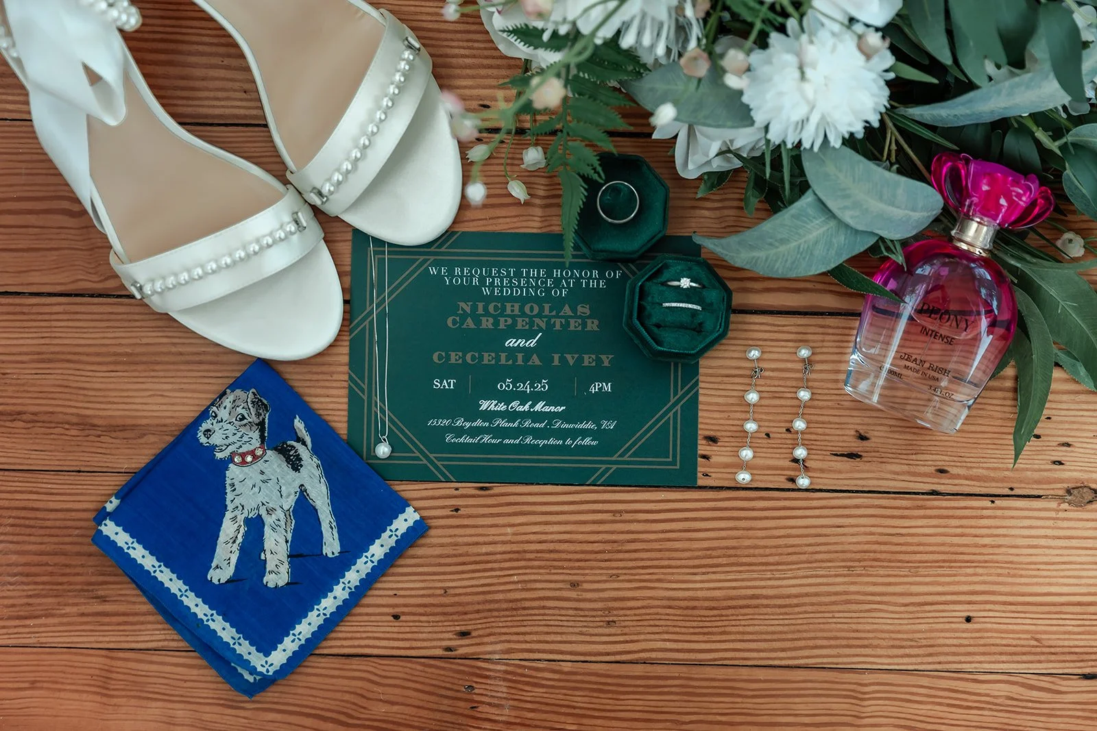 Wedding invitations, white shoes with pearl details, a blue napkin with a dog illustration, pearl earrings, a ring in a green holder, perfume bottle, and a bouquet of white flowers on a wooden surface.