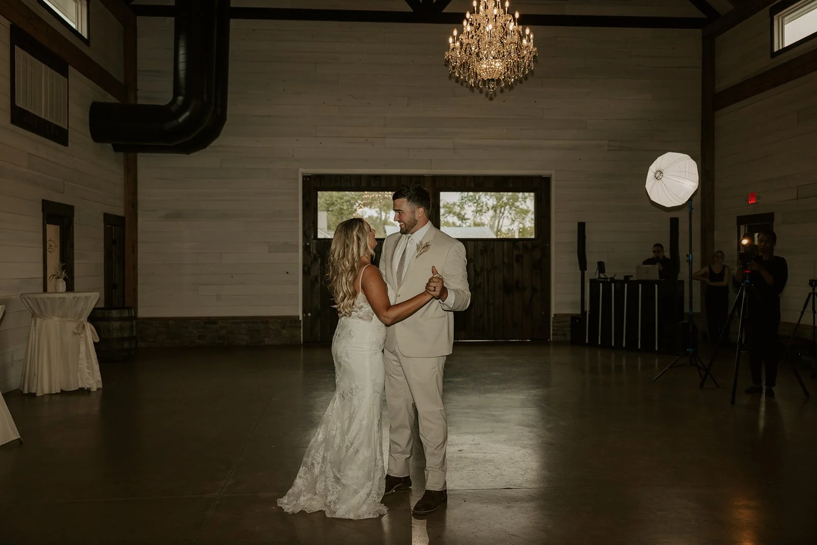 A bride and groom sharing a dance in a decorated reception hall with wooden walls, a chandelier, and dance floor lighting.
