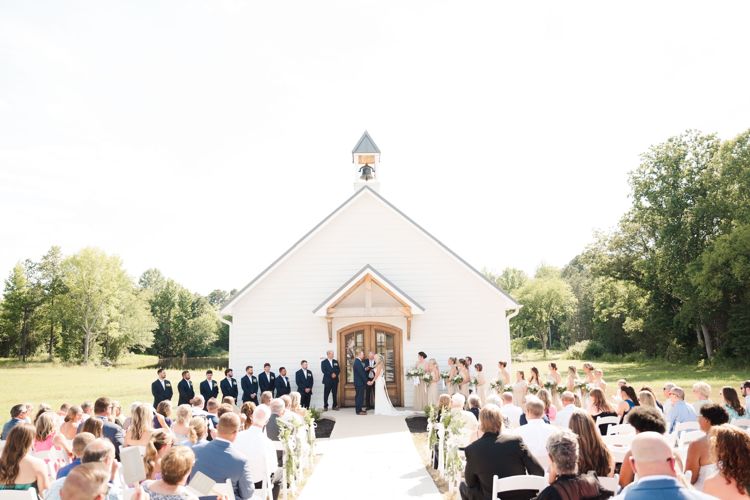Outdoor wedding ceremony in front of a white chapel with a seated guests and bridal party.